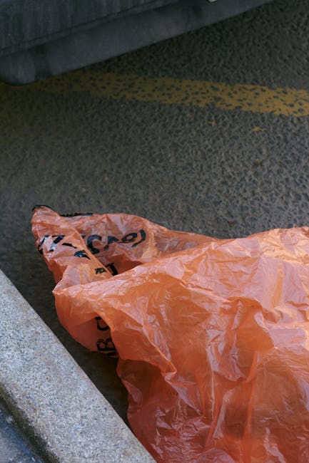 An orange plastic rubbish bag lies on a rough asphalt surface next to a concrete curb, partially resting on the pavement and partially on the road. The bag appears to be crumpled and contains waste, w