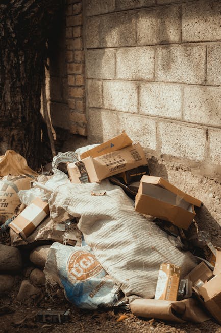 An orange plastic rubbish bag lies on a rough asphalt surface next to a concrete curb, partially resting on the pavement and partially on the road. The bag appears to be crumpled and contains waste, w