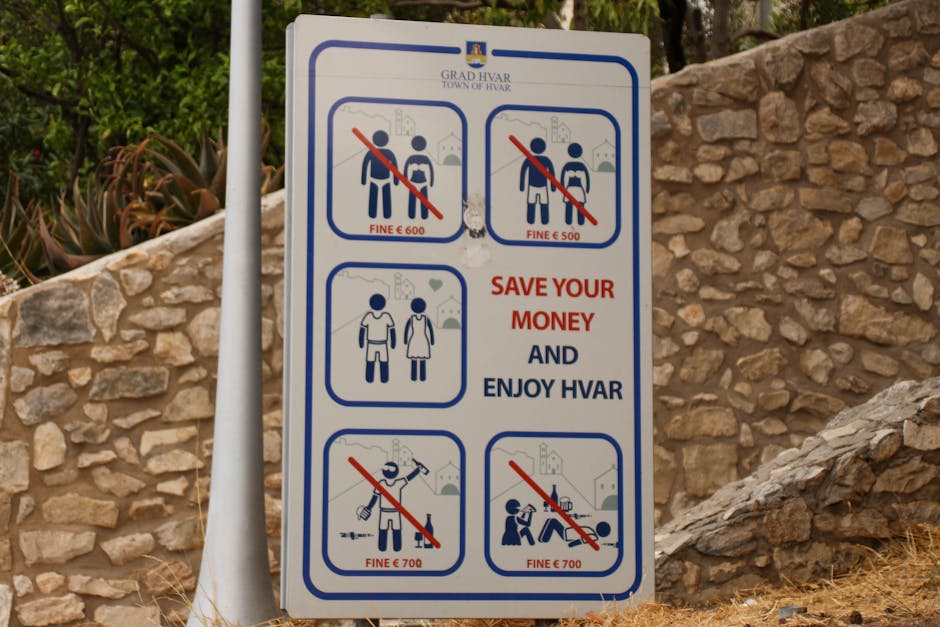 A vertical rectangular informational sign mounted on two white metal posts, situated outdoors against a backdrop of a stone wall and some greenery. The sign displays text and pictograms related to was