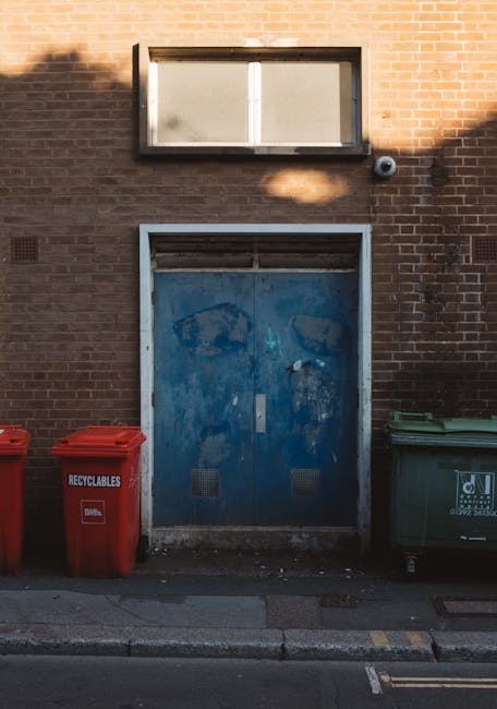A tall, bright blue plastic rubbish bin with a partially open lid, situated outdoors on a grassy area. Attached to the front of the bin is a small rectangular sticker with a jagged edge, bearing the t