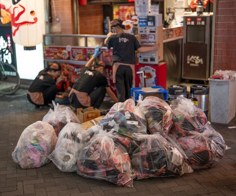 A clear view of a large pile of various types of discarded waste materials, including cardboard boxes, plastic containers, and metal objects, all loosely stacked outdoors on a paved surface. The waste